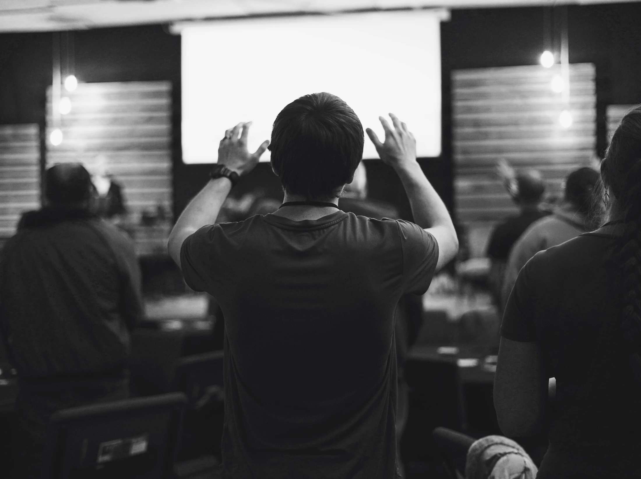 A black and white photo from behind a young man with his hands raised. He is in a crowd of people looking at a bright, blank screen at the front of a room.